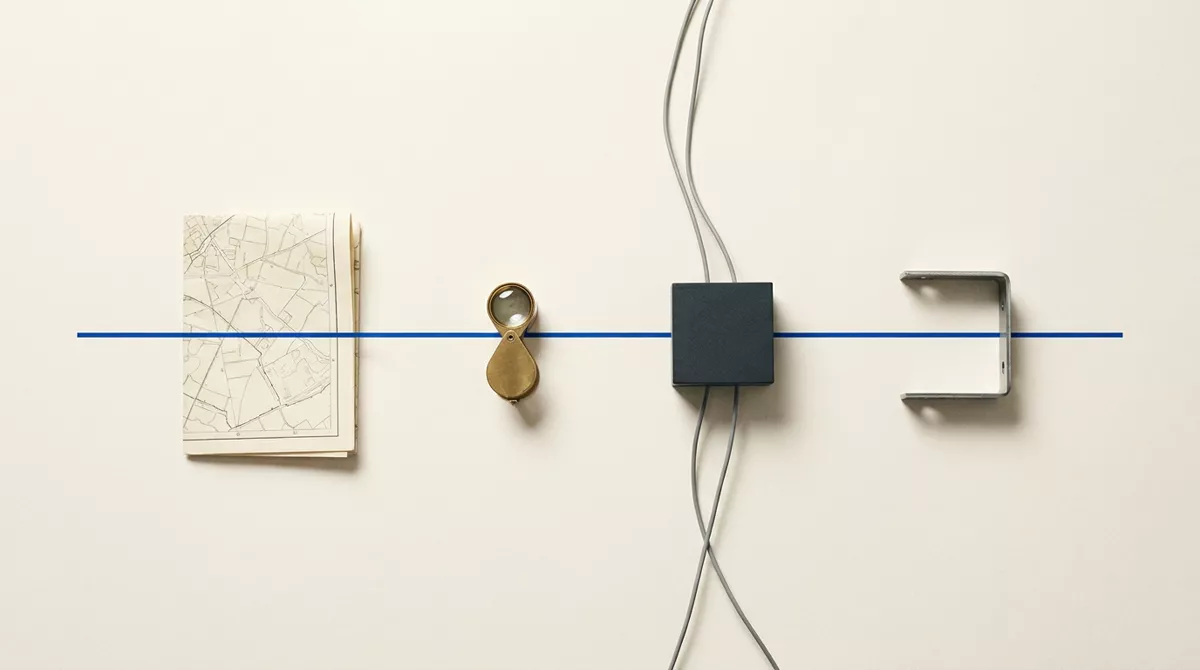 A folded map, a brass loupe, a charcoal block with grey cables and a steel bracket in a row, crossed by one blue line.